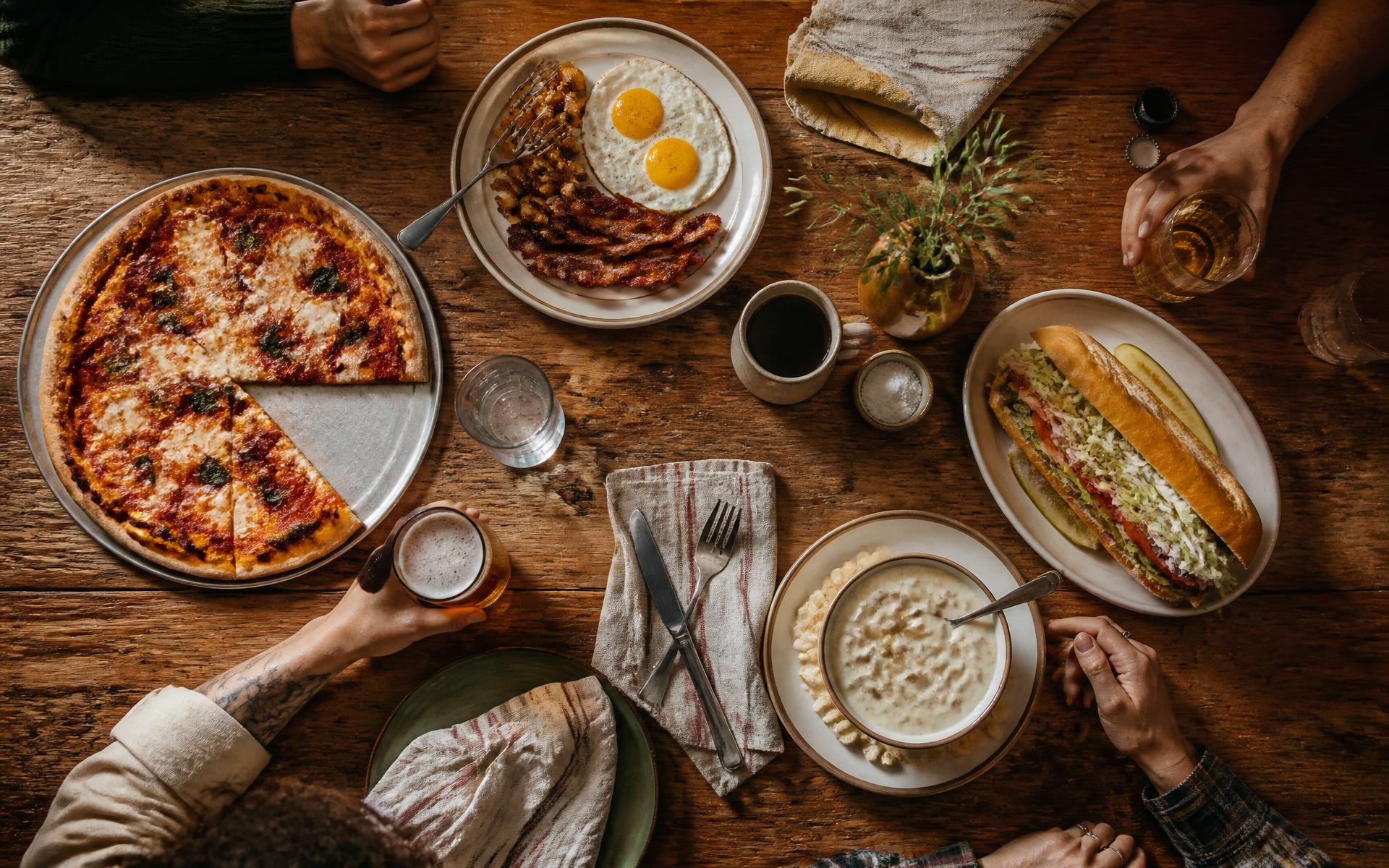 An overhead view of a long wooden table set with pizza, a breakfast plate, a sub and a bowl of chowder